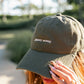 A woman outdoors wearing an olive green dad hat with 'planty aunty' embroidered in white on the front, holding the brim with her hand.