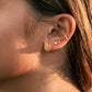 A close-up side profile of a woman's ear wearing a textured gold flower-shaped stud earring and a small white pearl stud, with warm sunlight and her hair in the background.