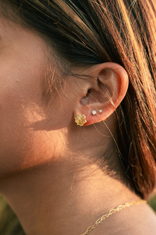 A close-up side profile of a woman's ear wearing a textured gold flower-shaped stud earring and a small white pearl stud, with warm sunlight and her hair in the background.